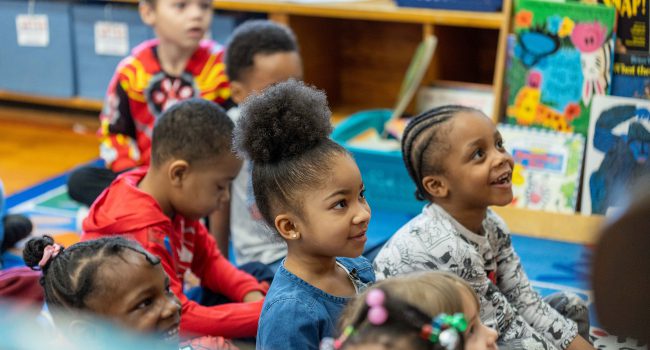 Young children sit on a rug and smile up at their teacher.