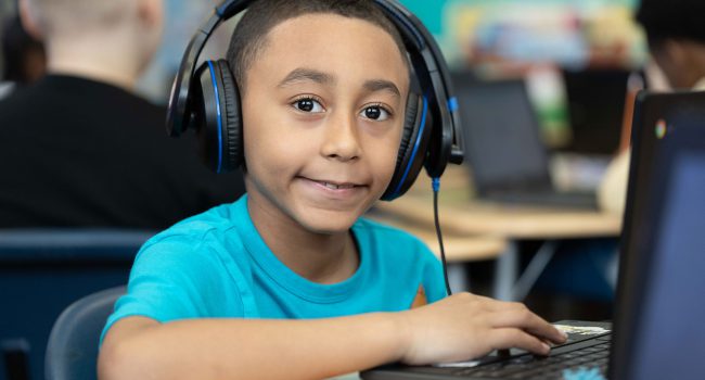 A student with headphones smiles as he uses a computer.