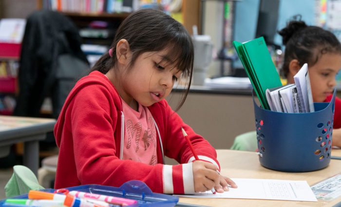 A young child in a classroom erasing her writing from a piece of paper.