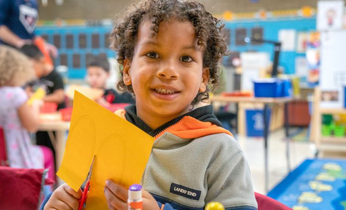A young child smiling as he cuts a piece of paper.