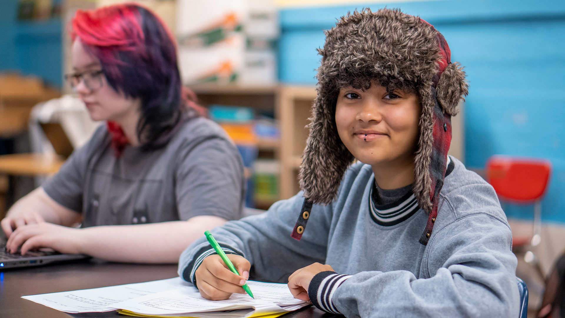 A smiling student wearing a winter hat smiles as they write.