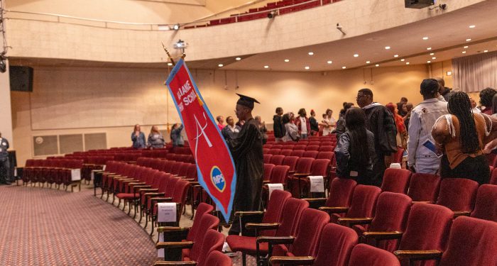 A graduate enters the auditorium holding the Alliance School banner.