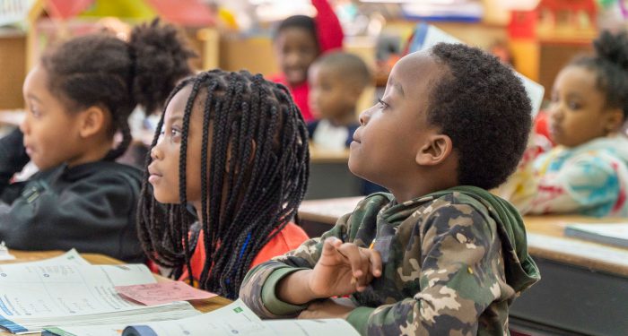 Young children in a classroom look to the front of the room.