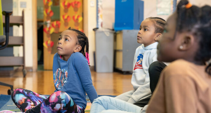 Young children in a classroom sit on the rug looking up a teacher.