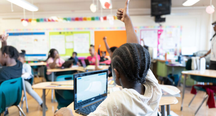 Children in a classroom, with many raising their hands.