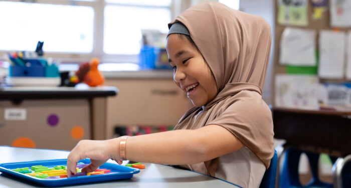 A young child in a hijab smiles as she adds magnetic letters to a tray in front of her.