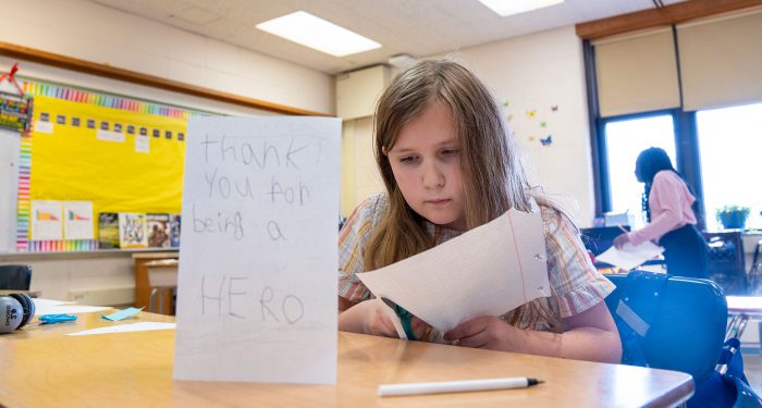 A child in a classroom cutting a piece of paper with a homemade card on the desk in front of her that reads "thank you for being a hero."