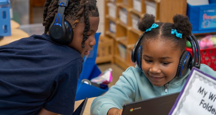 Two young children wearing headphones look at a computer screen.