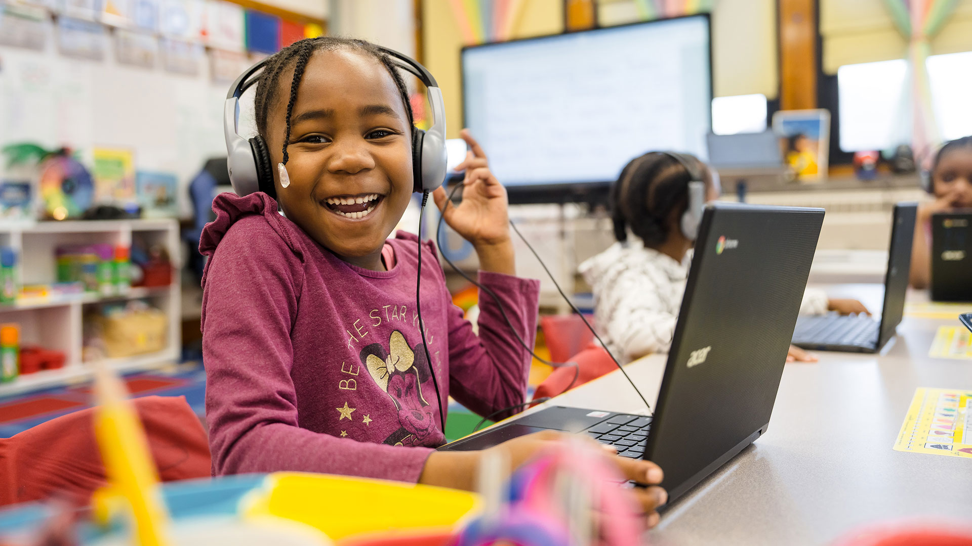 A young girl with headphones on and a laptop in front of her, smiling.