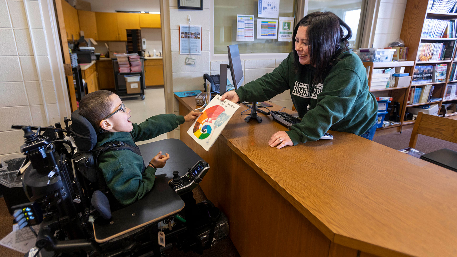 A librarian hands a picture book to a young boy in a motorized wheelchair.