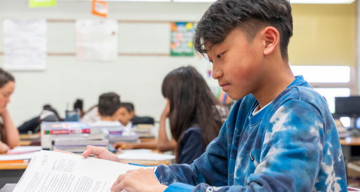 A boy in a classroom reading a textbook.