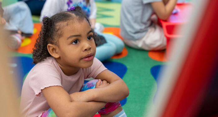 A young child sits on a colorful rug with her arms on her knees.