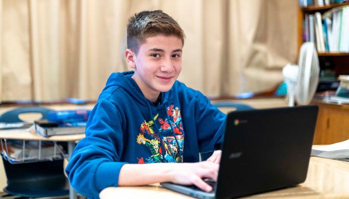 A boy with a superhero sweatshirt smiles as he uses a computer.