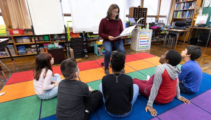 A teacher reads to her students as they sit on a colorful rug.