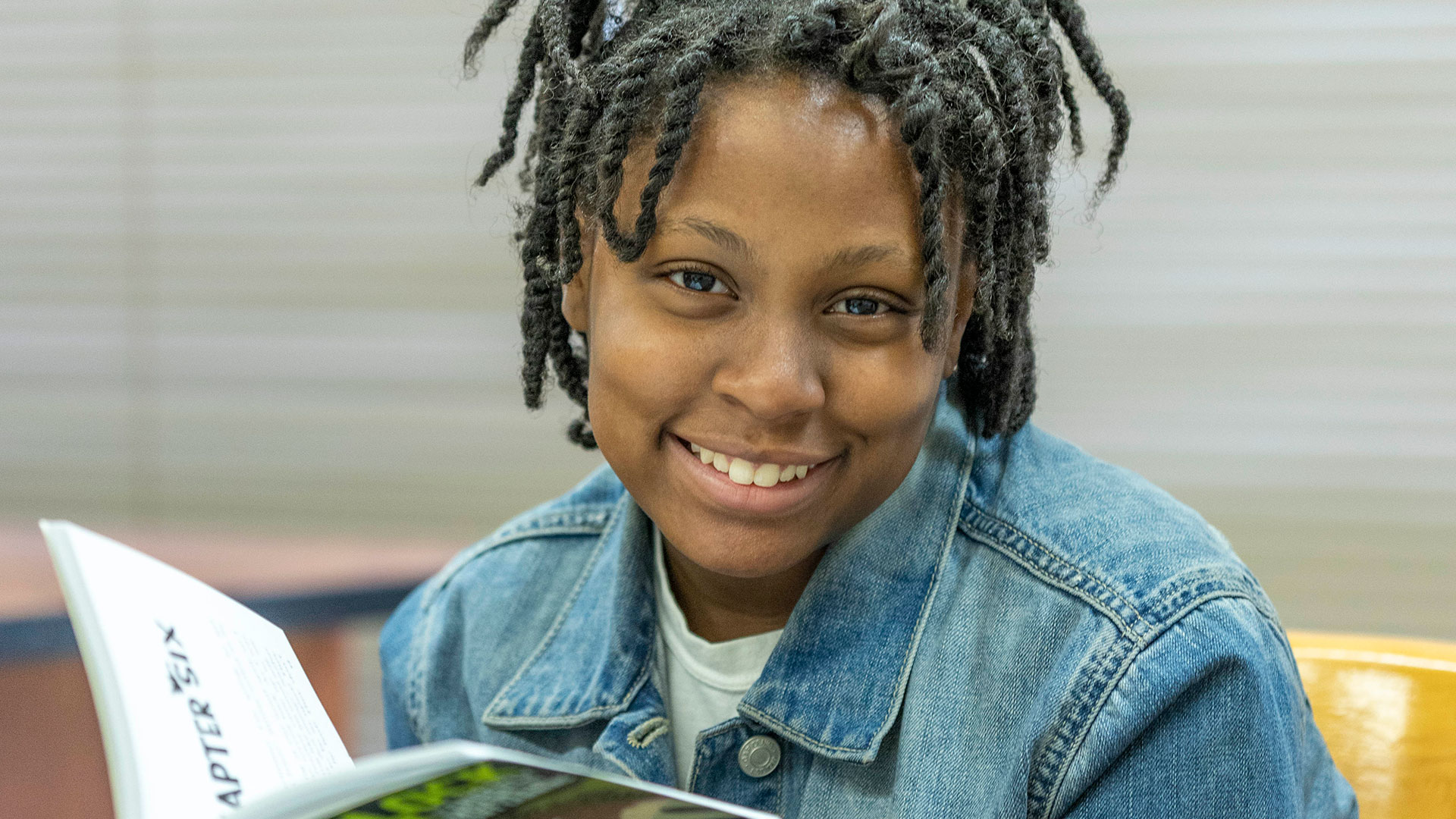 A girl reading a book, smiles at the camera.