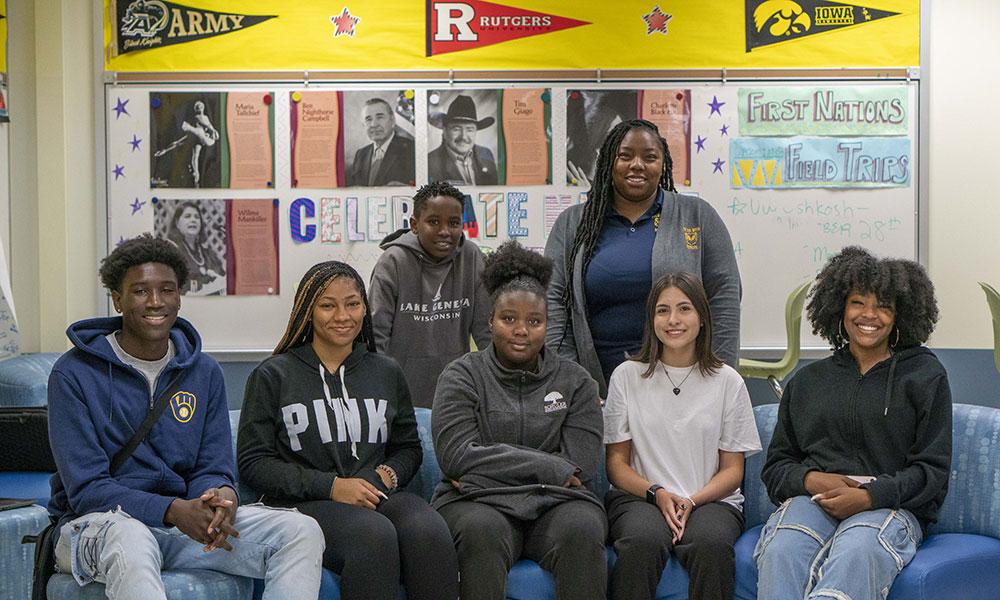 Six students sit on a couch with their teacher behind them.
