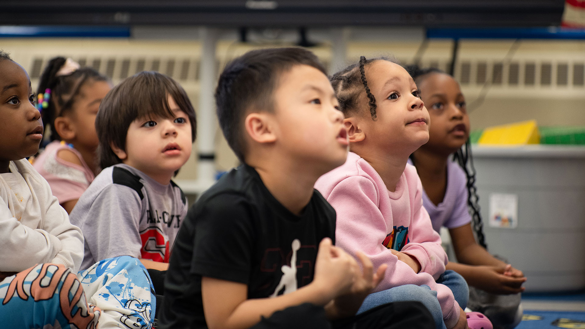 Young students sitting on a rug looking up at their teacher.