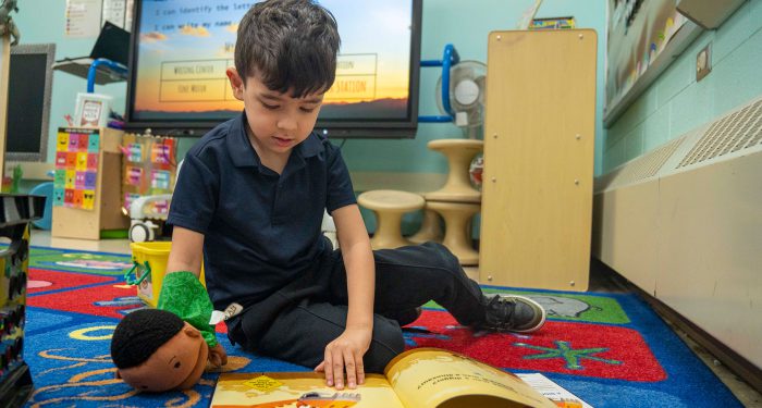A child sits on a carpet and looks at a picture book with a puppet on his hand.