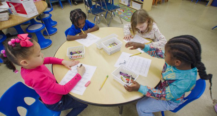 Overhead view of four young children at a table writing on worksheets.