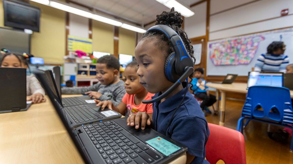 Young students sit at tables and use laptops.