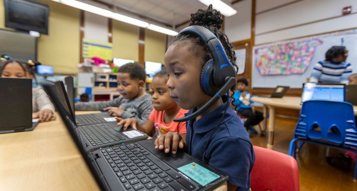 Young students sit at tables and use laptops.