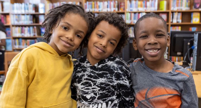 Three young students smile as they stand side-by-side in a library.