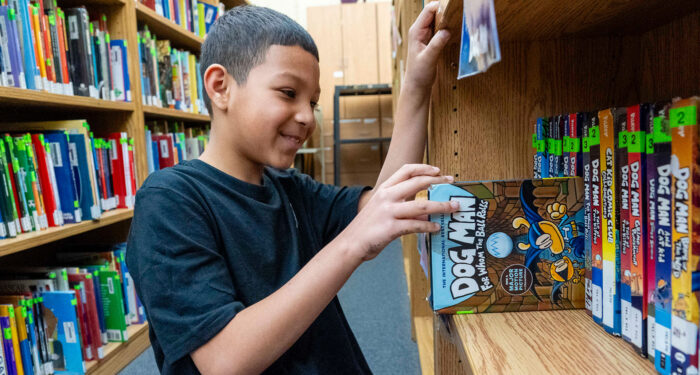 A smiling child in a library pulling a Dog Man book from the shelf.