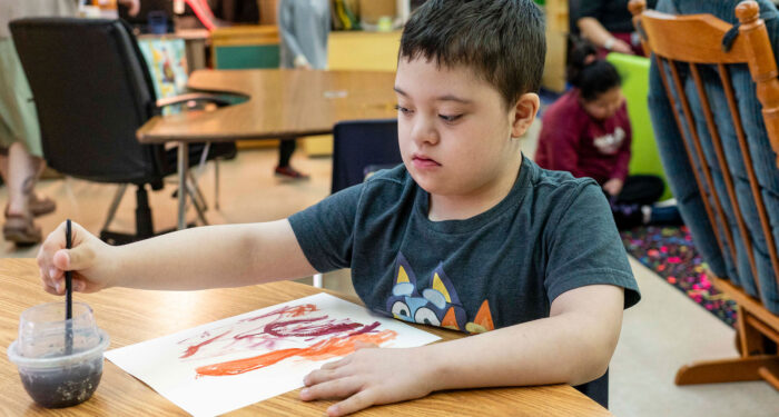 A child rinses their paintbrush in a clear cup of water with a colorful painting on the table in front of them.