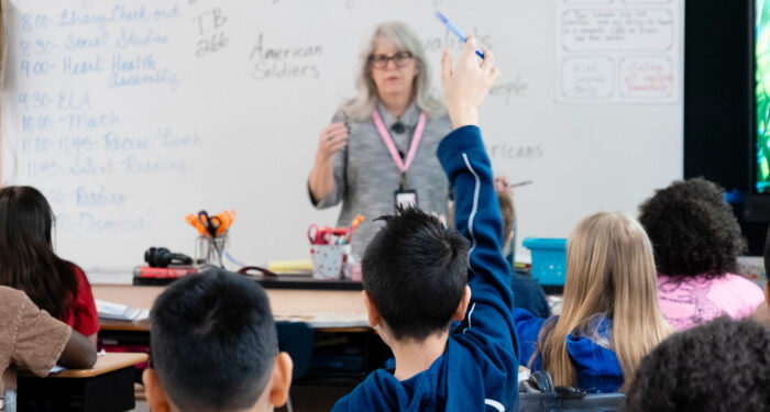 View from behind a classroom looking toward a teacher, with a child in the foreground raising his hand.
