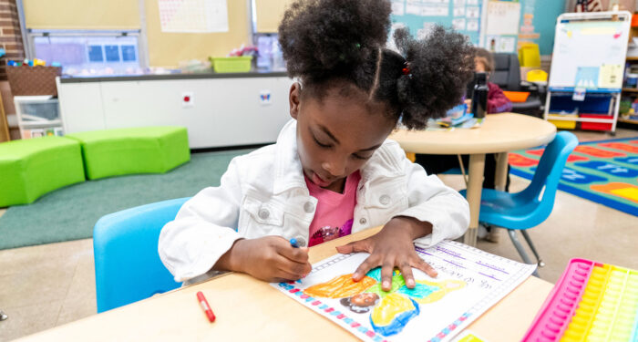 A young child in a classroom with colorful furniture works on a drawing.