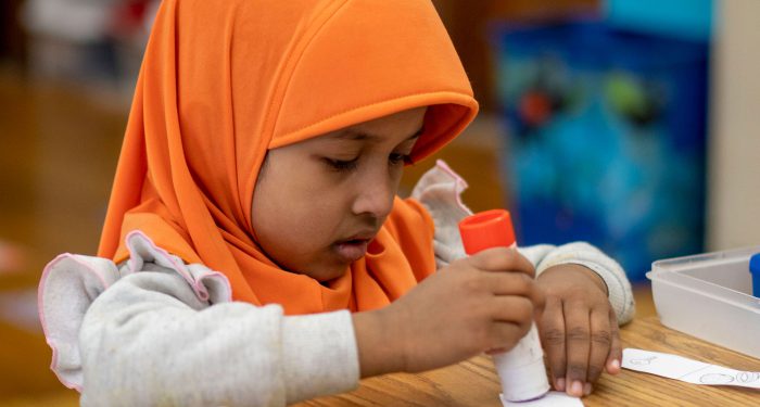 A young girl with a colorful hijab applies glue to a strip of paper.