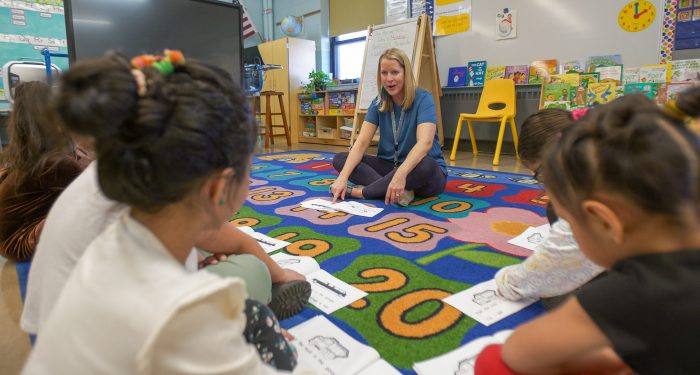 A teacher sits on a colorful rug with her students.