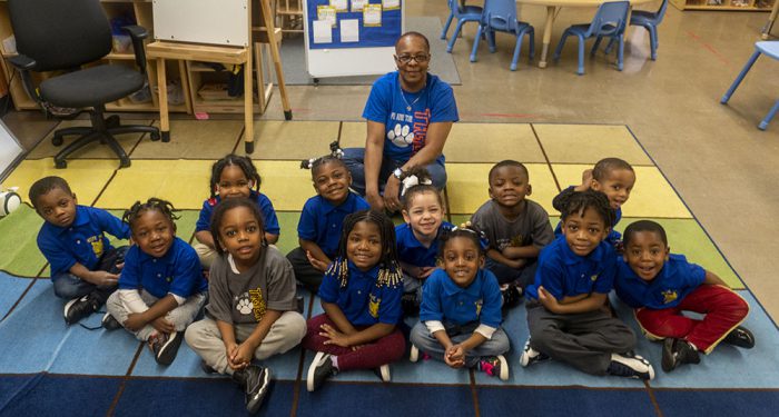 Young students sit on a rug with their teacher behind
