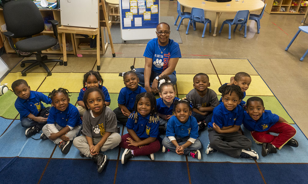 Young students sit on a rug with their teacher behind