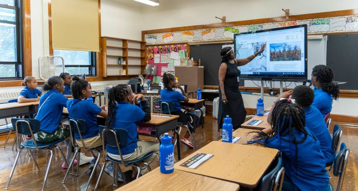 Students in a classroom watch as the teacher points to a large screen.