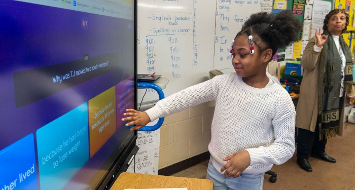 A student selects an answer on a large touchscreen while the teacher looks on.