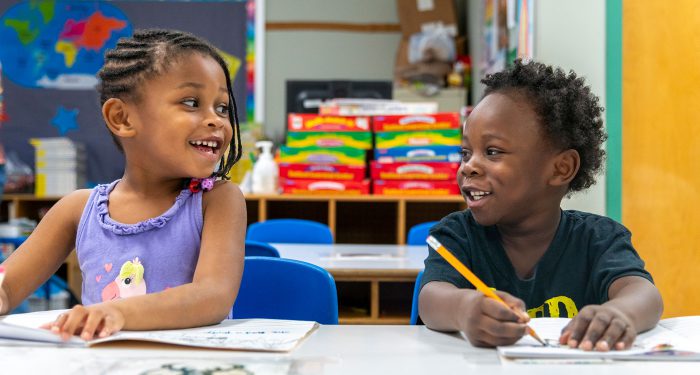 Two young children in a classroom smiling at one another.