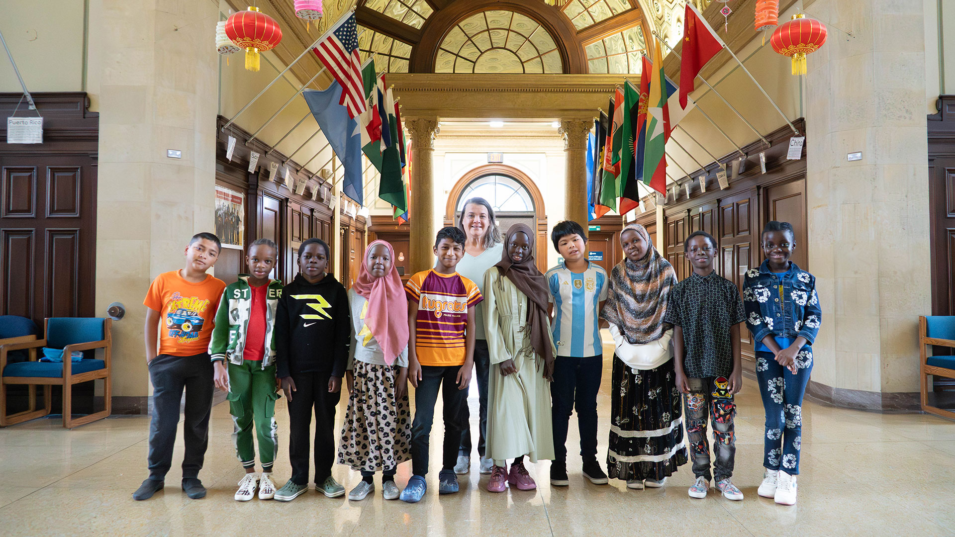 Young children and a woman in a hallway with flags from different countries.
