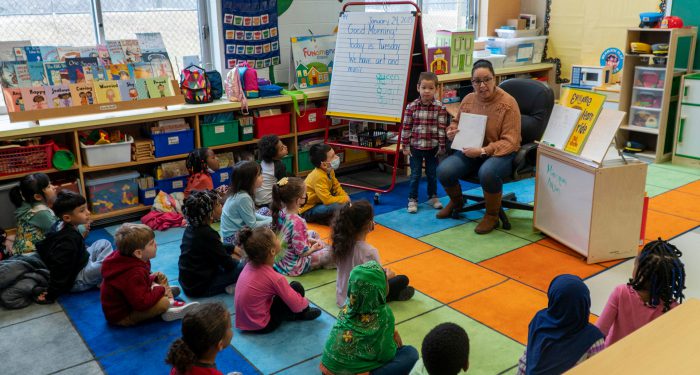Young students sit on a colorful rug and look up at the teacher.
