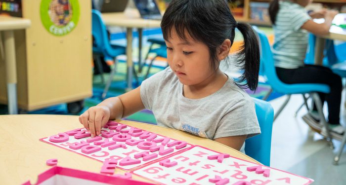 A young girl sits at a table and matches wooden letters to a lettered placemat.