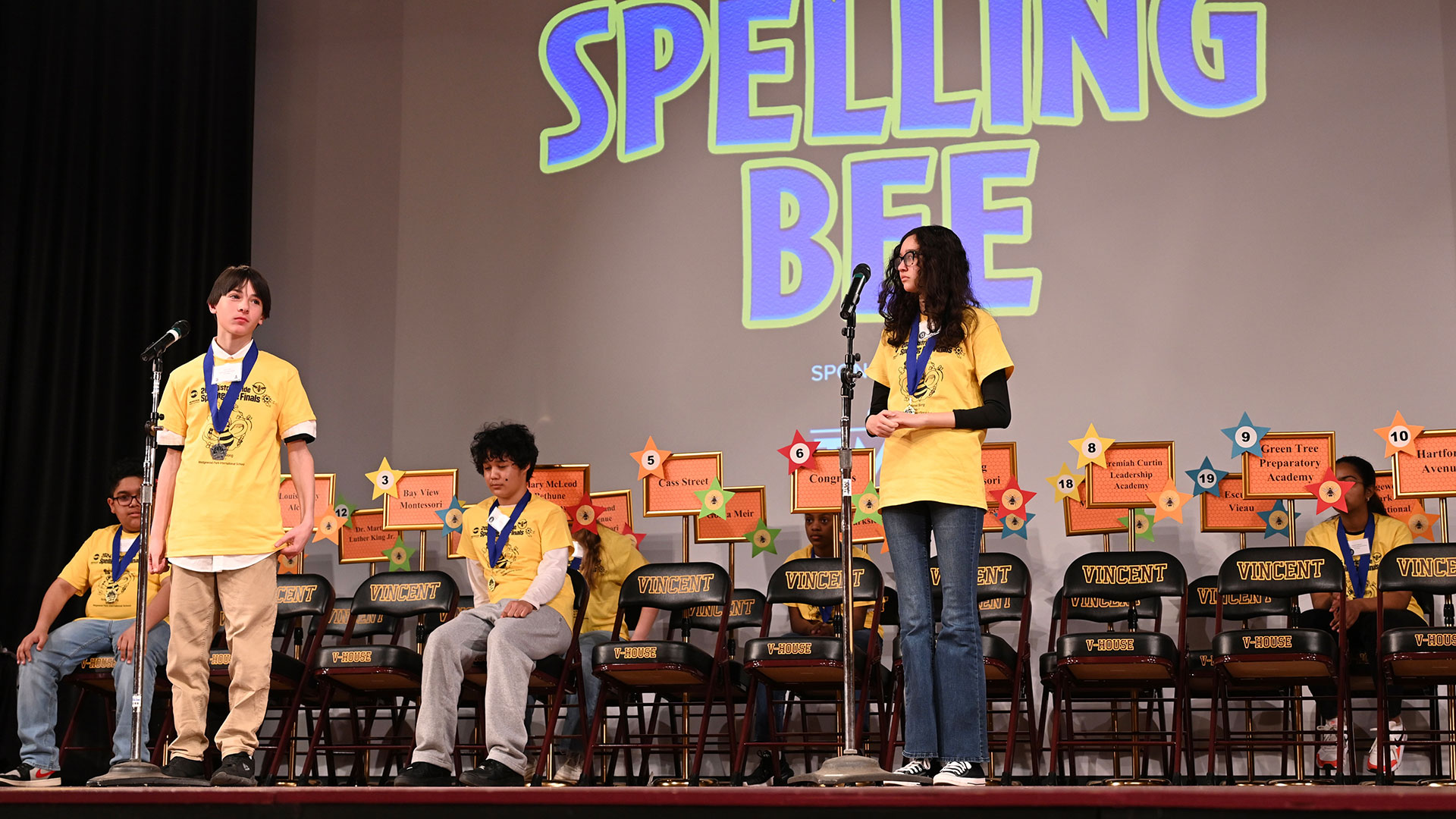 Ellen on stage, watching as a fellow student spells his word.