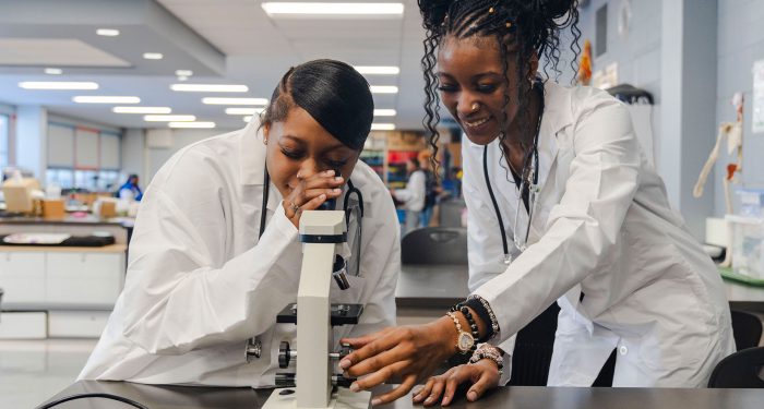 Students in lab coats look through a microscope.
