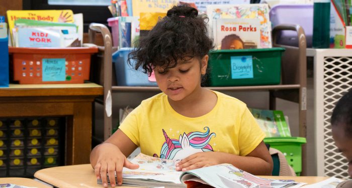 A young girl in a classroom reading a book.