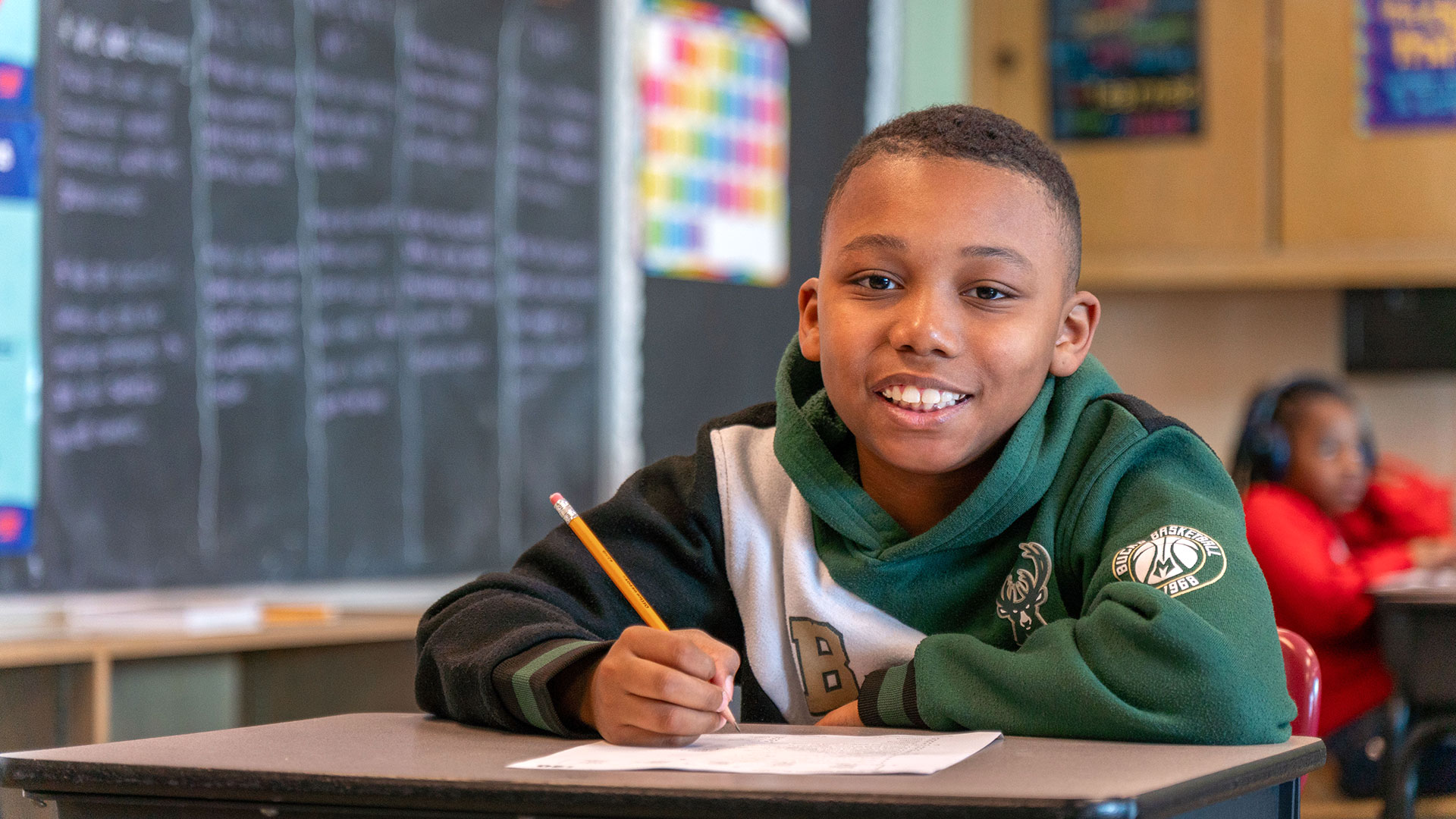 A boy in a classroom prepares to write as he smiles at the camera.