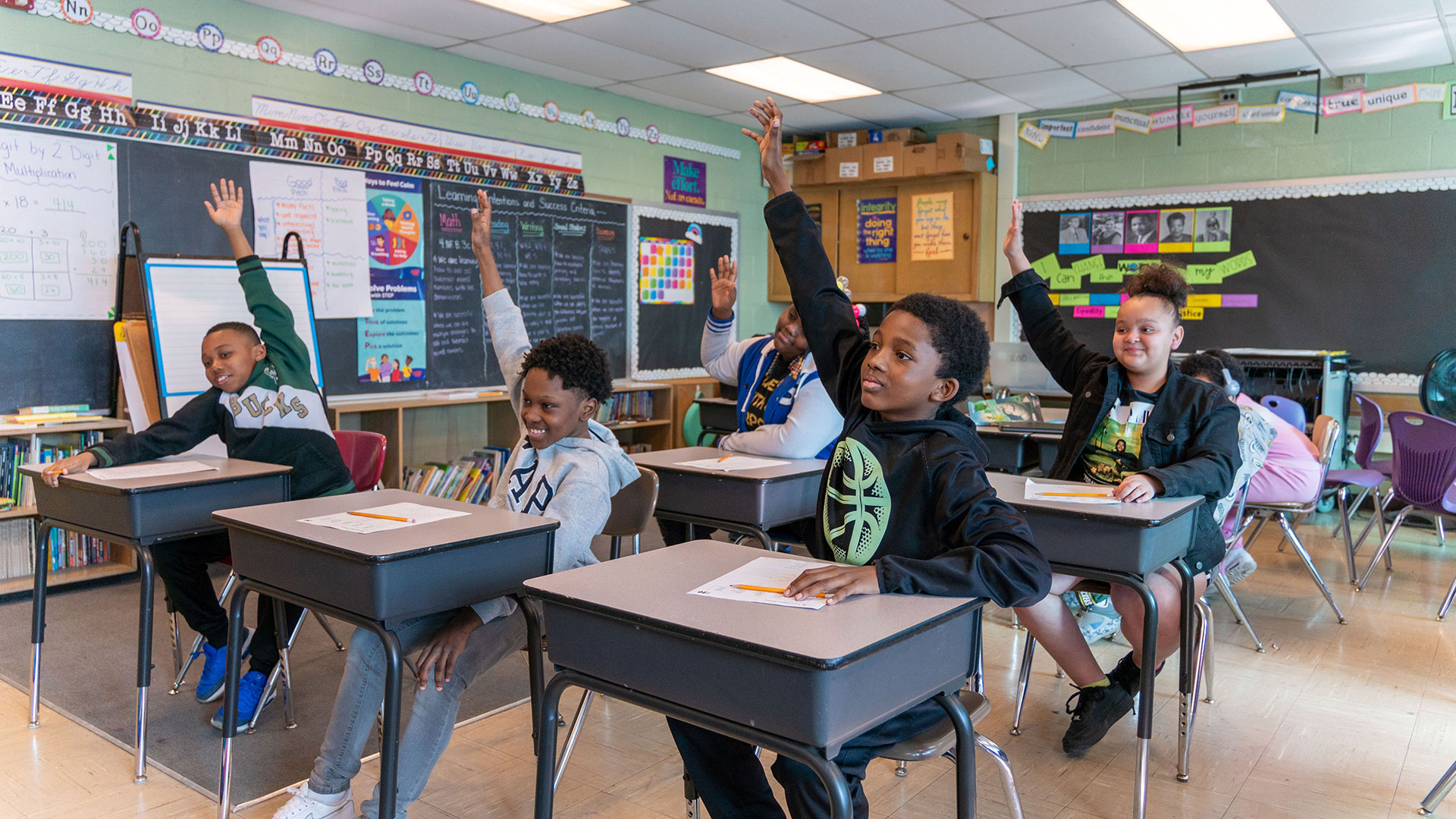 Students raising their hands to answer a question as they sit at desks in a classroom.