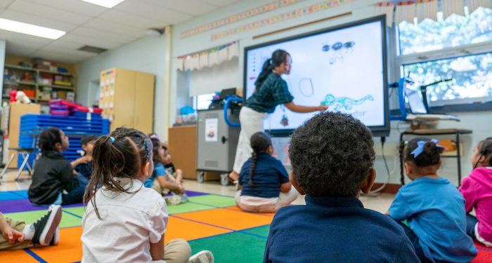 Young students sit on a colorful rug and look up at their teacher.