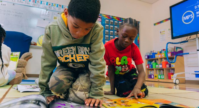Two young students sit on the floor and look at picture books.
