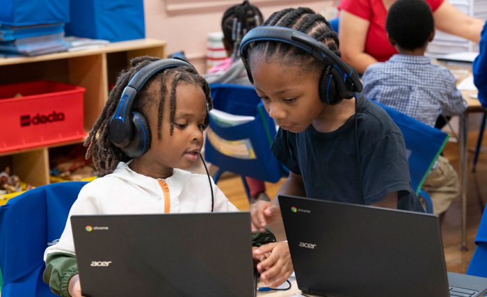 Two young children in a classroom helping one another with their computers.