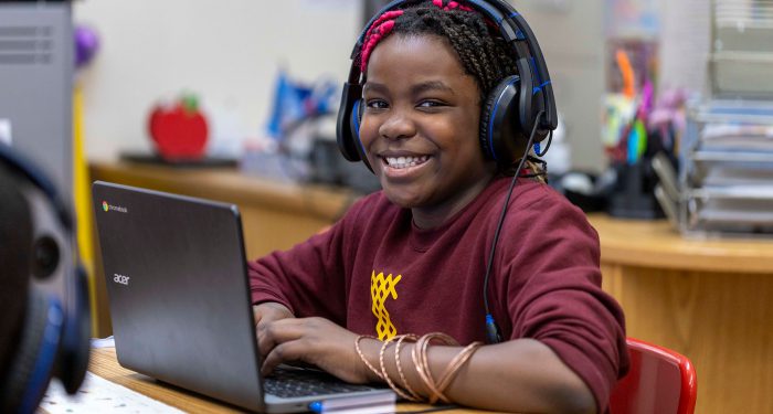 A girl with headphones smiles as she types on her laptop.