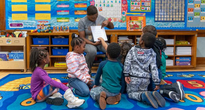Young students sit on a colorful rug at the foot of their teacher as she shows them a diagram.